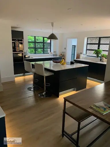 Dark kitchen island with plinth lighting and bar stools in an open-plan room with black steel-framed windows