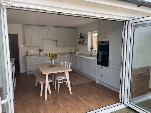 Light grey shaker kitchen with built-in ovens and a dining table, seen through open bifold doors from the garden