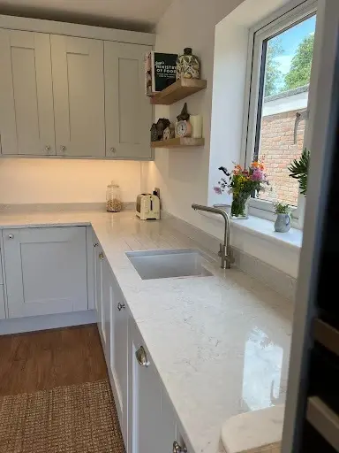 Light grey shaker kitchen with an undermount sink, white quartz worktops and floating oak shelves by the window