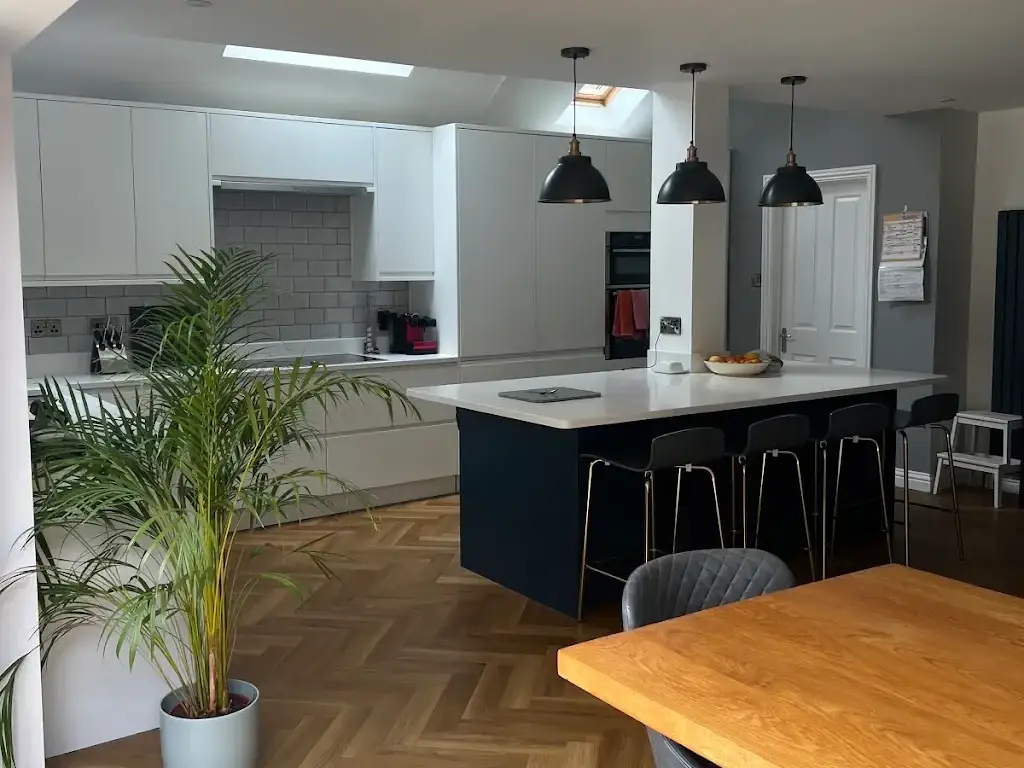 Open-plan kitchen with pale grey handleless units, a navy island with a white quartz top and bar stools, three black pendant lights, roof lights and herringbone flooring