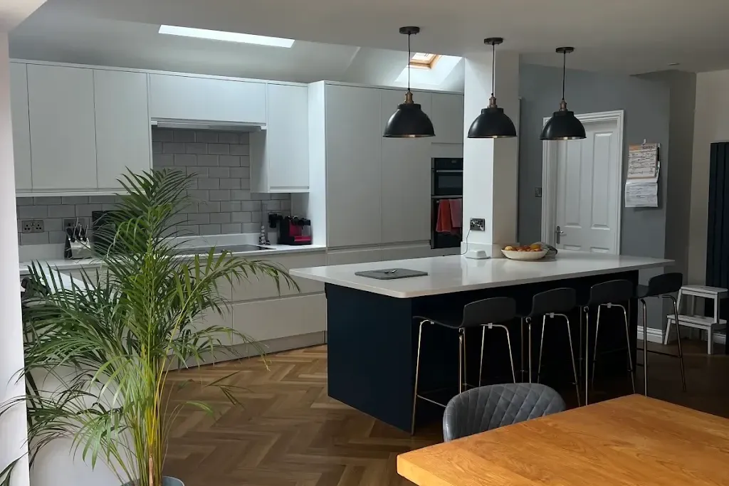 Open-plan kitchen with pale grey handleless units, a navy island with bar stools, three black pendant lights, roof lights and a herringbone floor
