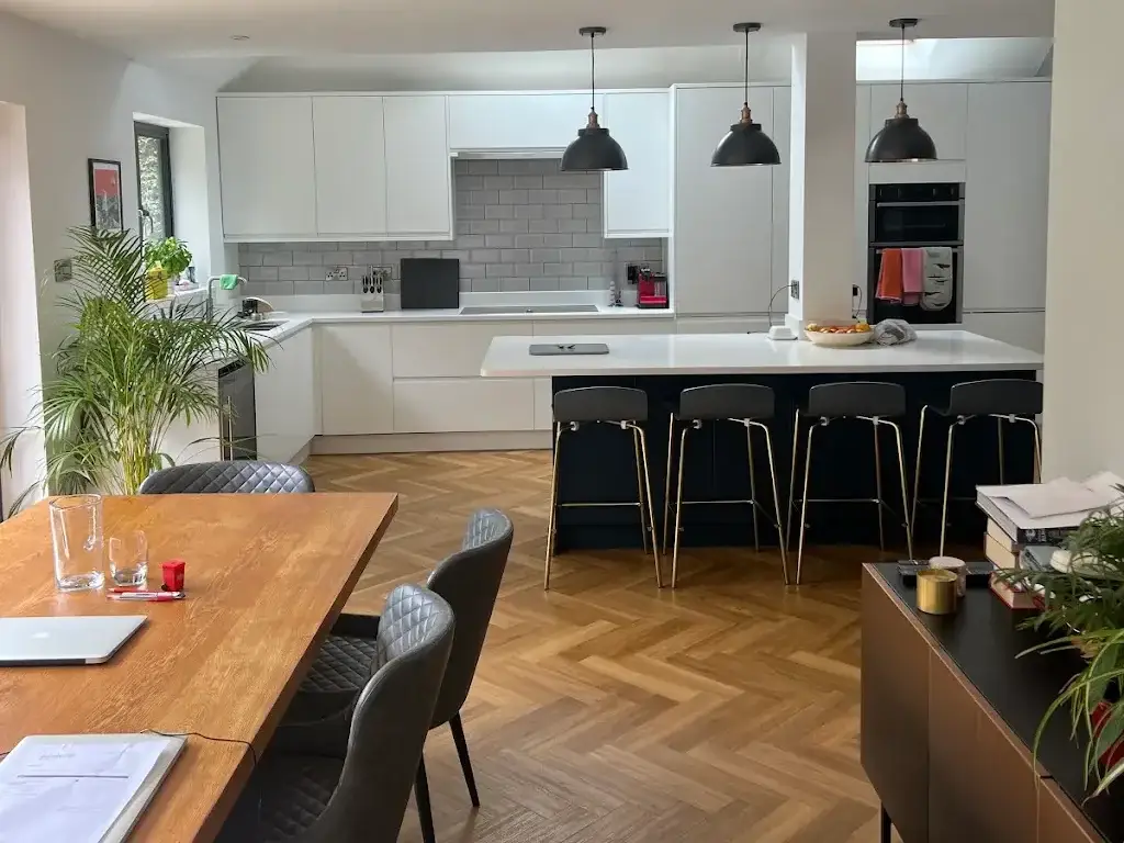 Finished open-plan kitchen with white handleless units, a navy island with four bar stools, three black pendant lights and herringbone flooring