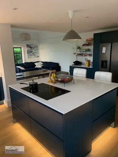 Navy handleless kitchen island with a white quartz top, induction hob and warm plinth lighting, open to a living area beyond