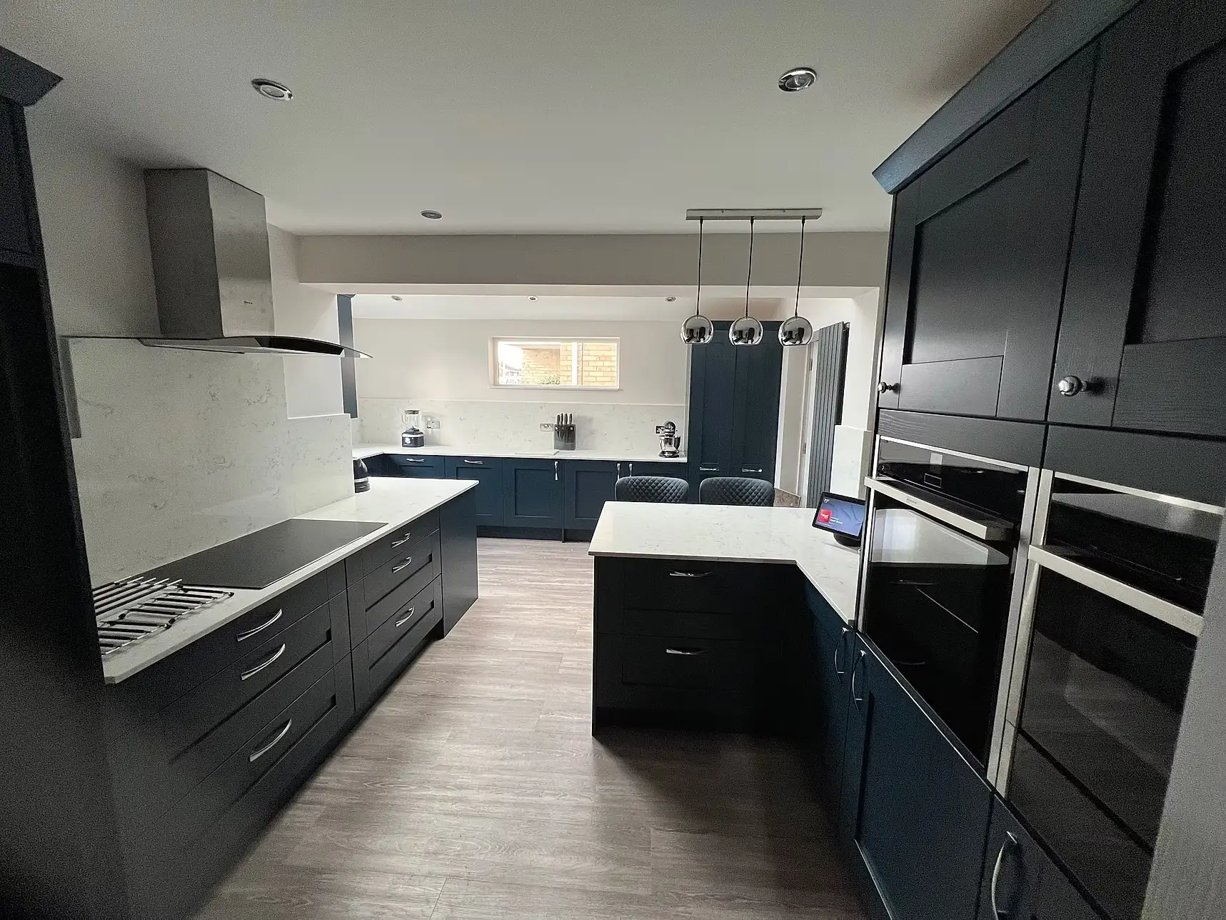 Finished navy shaker kitchen with quartz worktops, a peninsula, extractor hood, built-in double ovens and pendant lighting over a dining area beyond
