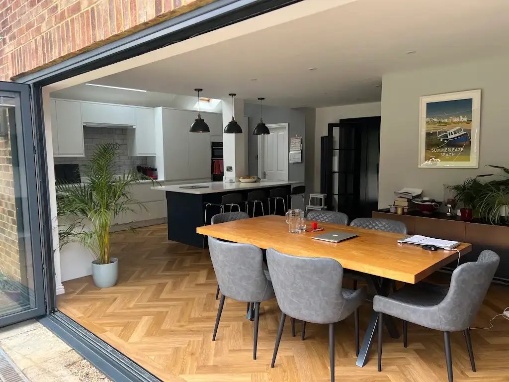 Open-plan kitchen and dining room seen through opened aluminium bifold doors, with herringbone oak flooring, a navy island and pendant lighting