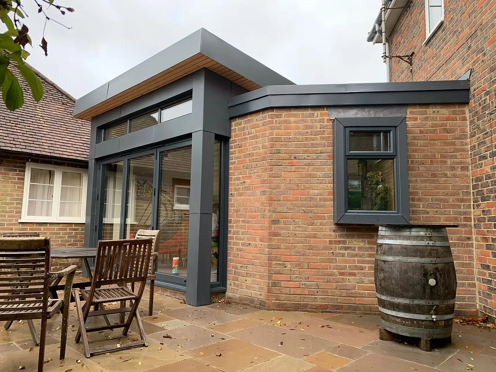 Completed single storey rear extension with a flat roof, dark grey aluminium glazing, brick flank wall and timber-lined soffit, opening onto a sandstone patio