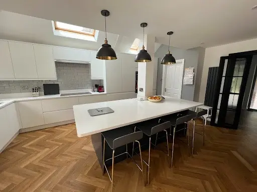White handleless kitchen with a large quartz island, bar stools, three black pendant lights and herringbone flooring under roof lights